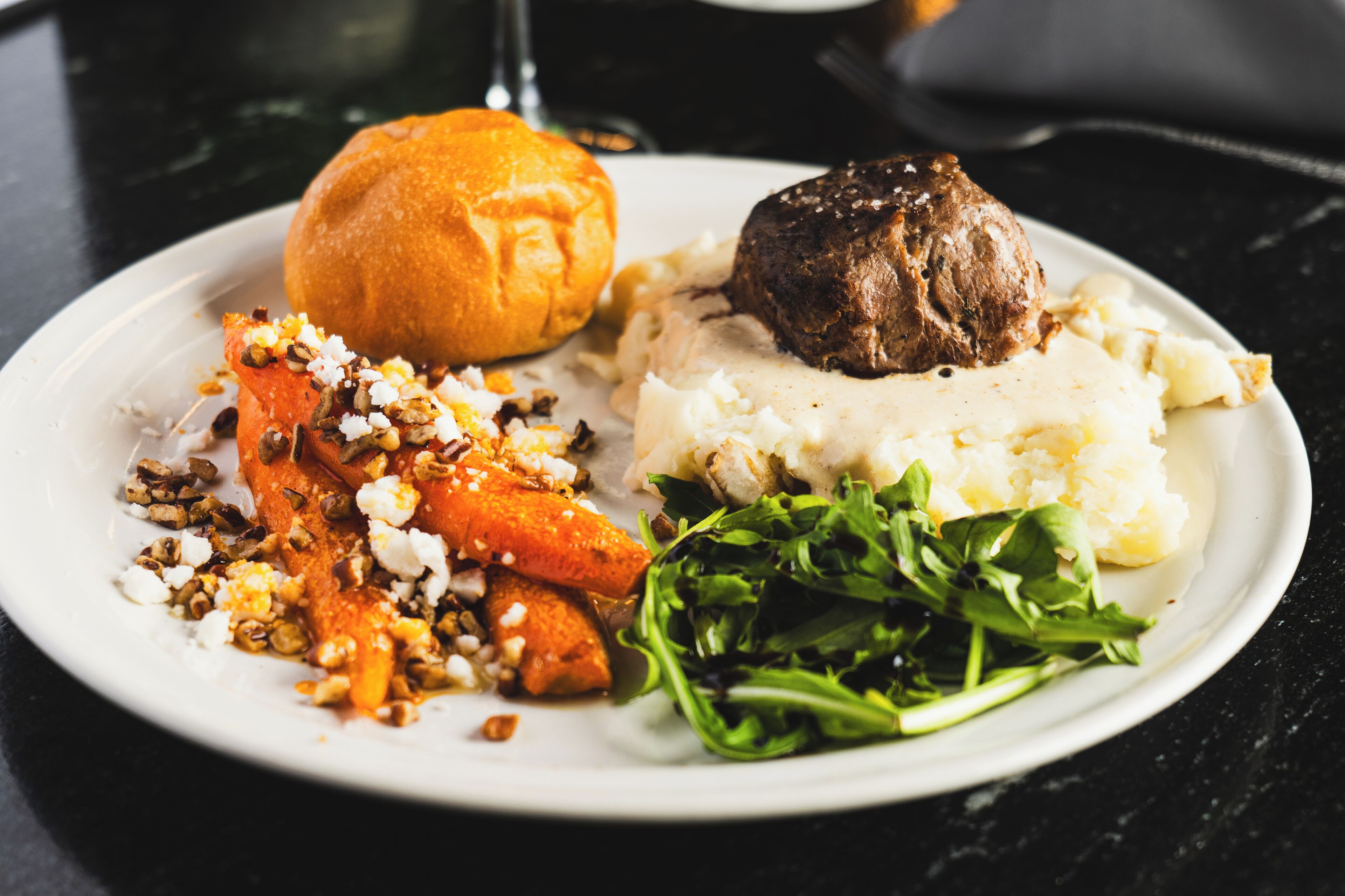 A plate with a meat patty, mashed potatoes, a roll, roasted carrots, and a side salad.