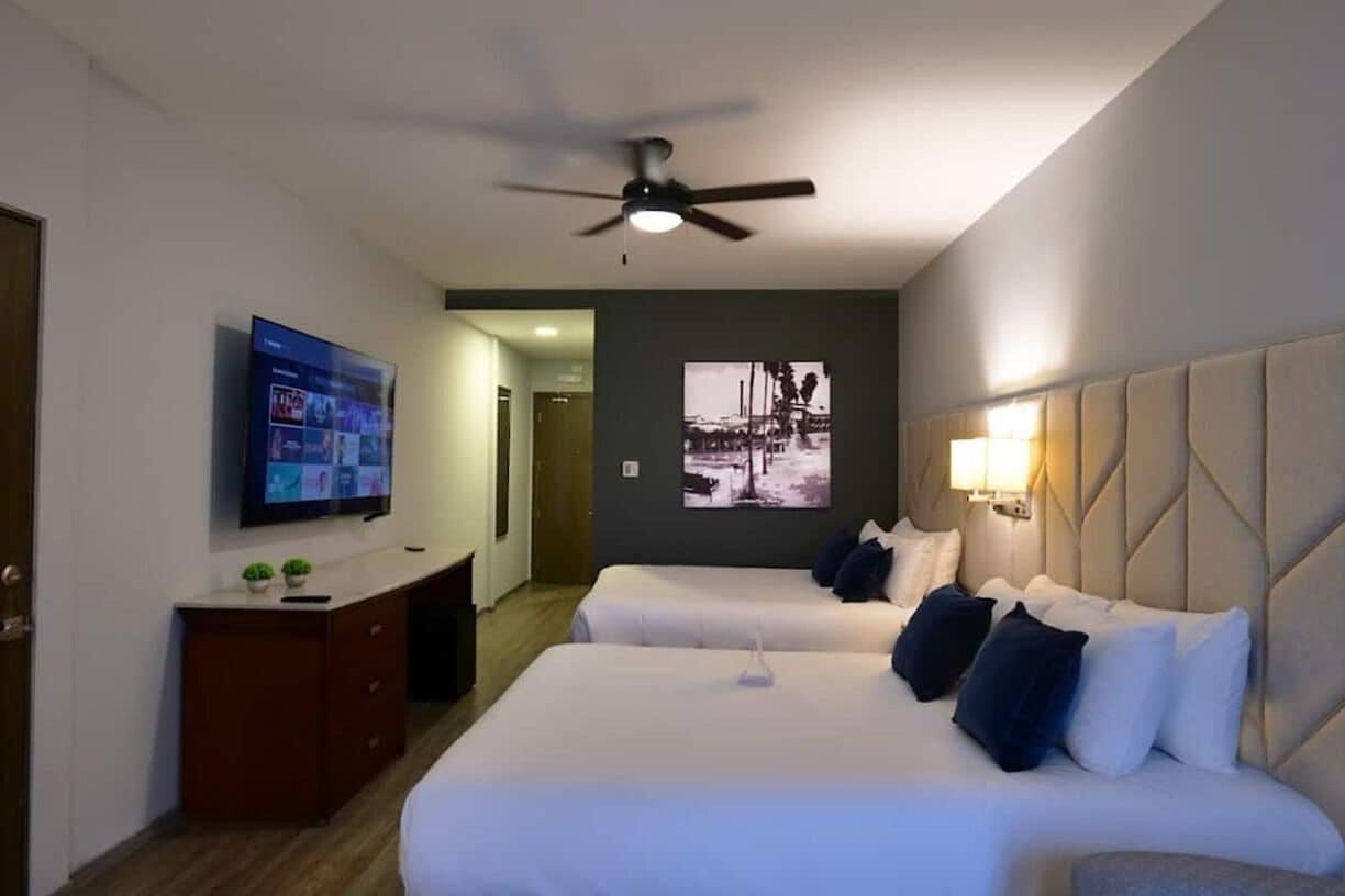 Habitación de hotel con dos camas, un televisor, ventilador de techo y un cuadro colgado en la pared.