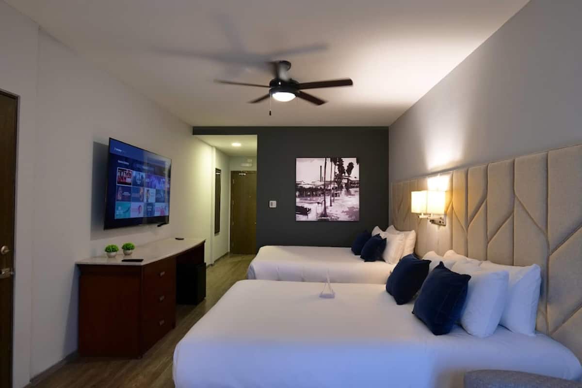 A hotel room with two beds, a TV, a ceiling fan, a desk, and a wall-mounted picture.
