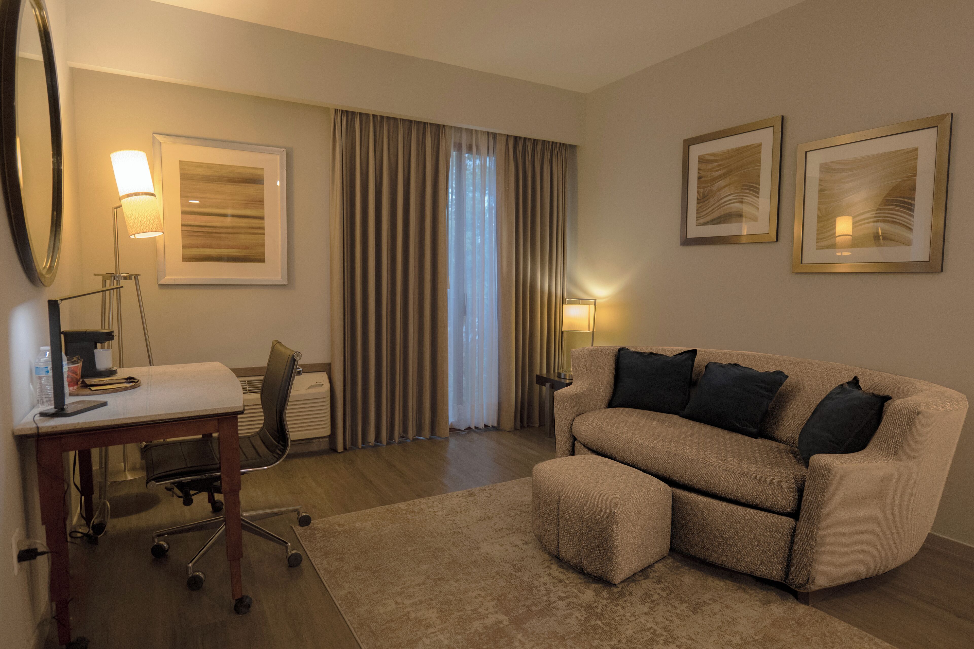 A hotel room with a desk, chair, sofa, and two framed artworks on the wall.