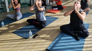 Una clase de yoga en una habitación moderna con ventanas amplias, suelos de madera y ventiladores de techo.