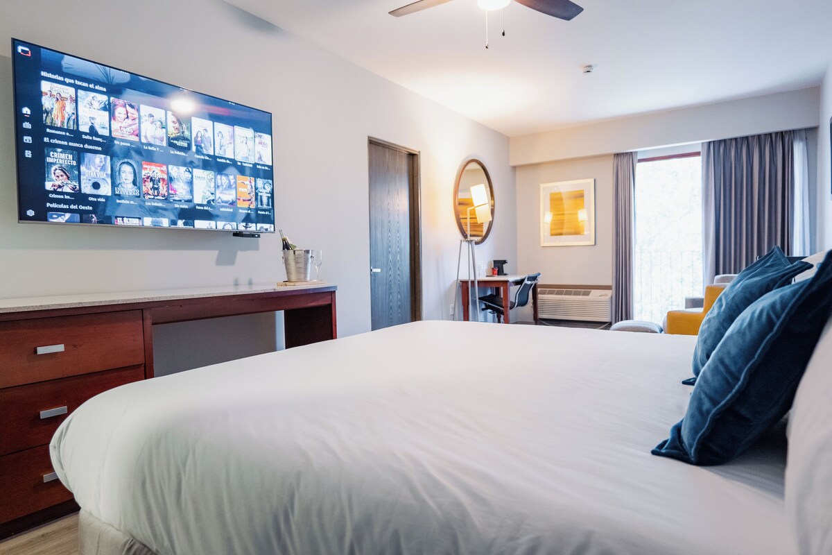 A hotel room with a large flat-screen TV displaying movie options, a bed with white linens, a desk with a lamp, and a window with curtains.