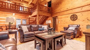 A spacious log cabin interior with a large leather sofa set, a dining area, and a staircase leading to an upper level. - The Log House at Twin Brook (Gap)