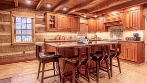 A rustic kitchen with wooden cabinets, a large island with bar stools, and a stone backsplash. - The Log House at Twin Brook (Gap)