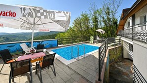 Zona de piscina con vista a las montañas, una mesa con sillas y sombrillas.