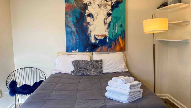 A bedroom with a large painting of a cow, a bed with a grey duvet, a black chair, and a lamp on a bedside table.