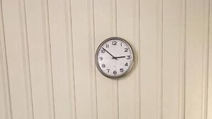 Une petite table blanche à deux places, surmontée d’une horloge ronde, placée devant un mur lambrissé et sur un plancher en bois.