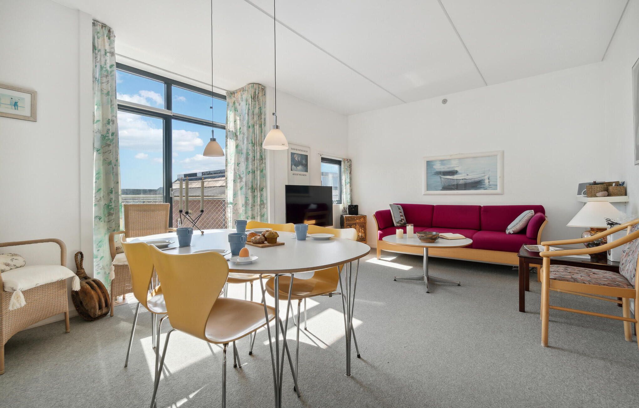 Ein modernes Wohnzimmer mit einem runden Esstisch, gelben Stühlen, einem roten Sofa und einem großen Fenster mit Vorhängen.