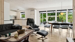 Un salon moderne avec un canapé, une table basse et un pouf, un coin repas avec des chaises, et une cuisine en arrière-plan.