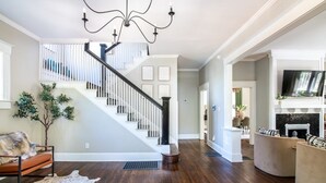 Un salon spacieux doté d’un plancher en bois, d’un escalier avec des rampes blanches, d’un lustre et d’une cheminée.