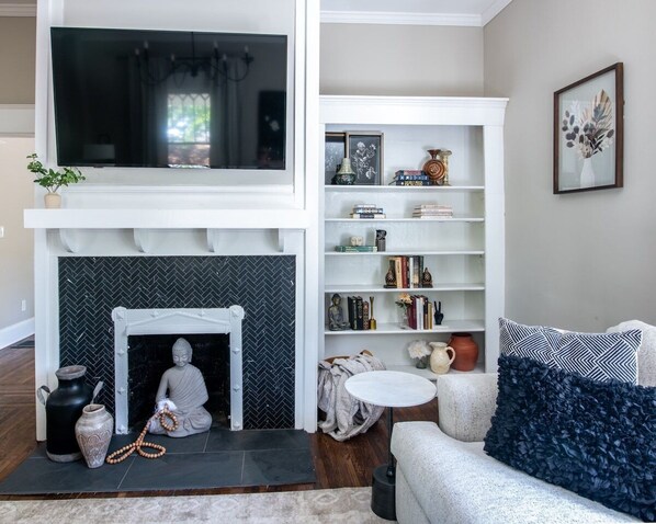 A living room with a fireplace, a television mounted above it, a bookshelf, and a sofa with a patterned cushion. - ✧Elmsgate✧ - A historic designer dream home w/ family friendly amenities , cozy porch&games near DT (Birmingham)