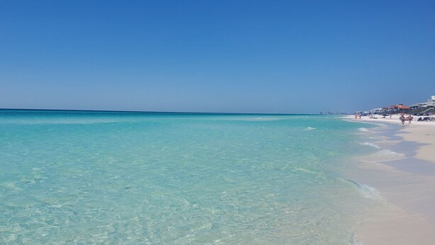 Une plage avec une eau turquoise et limpide, un rivage sablonneux et des bâtiments en arrière-plan.