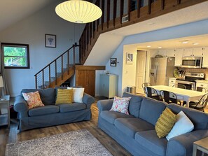 A spacious living room with two grey sofas, a wooden staircase, and a kitchen area with white cabinets and stainless steel appliances.
