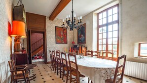 A dining area with a large round table, wooden chairs, and a chandelier. There is a staircase leading to an upper level, and artwork on the walls. - Demeure Historique - Village D'agon - 800m de la Plage (Agon-Coutainville)