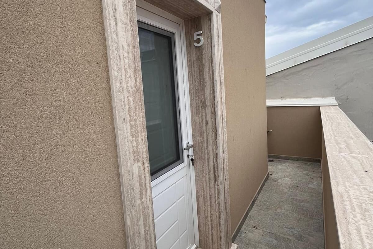 Un edificio con fachada color crema, una puerta blanca con ventanal de vidrio y un pequeño balcón con piso de concreto.
