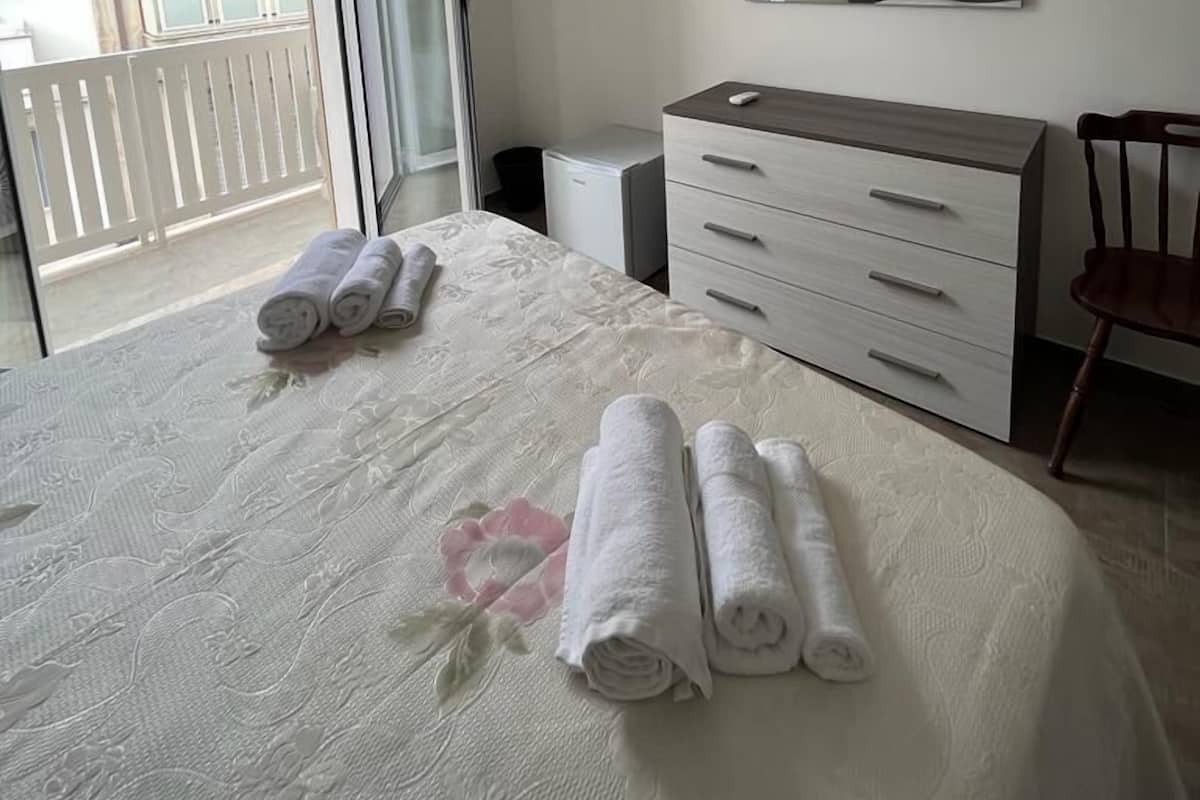 Un dormitorio con una cama, cómoda, silla, televisión y balcón.