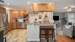 A modern kitchen with wooden cabinets, a stainless steel refrigerator, and a central island with stools. - Poolside Playhouse: Hot Tub, Firepit & Games (Stevensville)