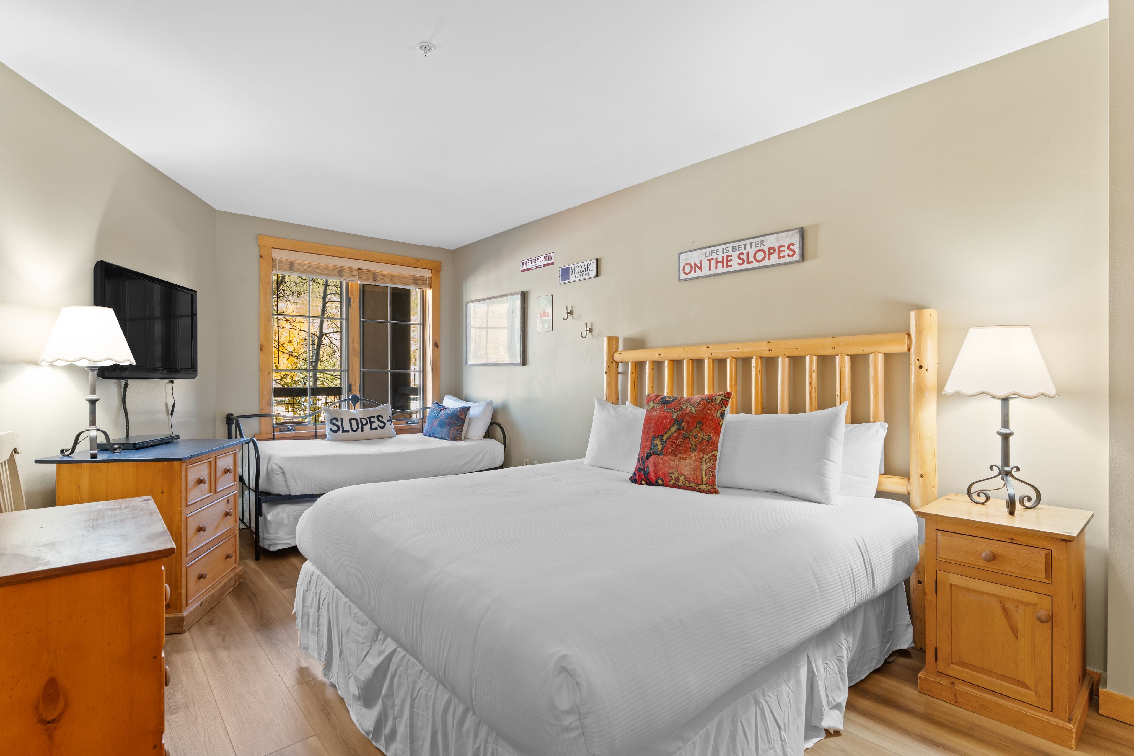 Une chambre avec deux lits, une tête de lit en bois, des tables de chevet avec des lampes, une commode, un téléviseur et une fenêtre donnant sur les arbres.