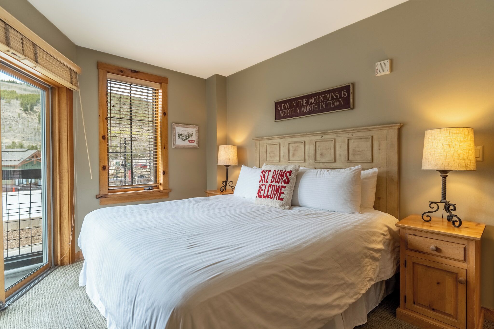 A neatly made bed with white linens and a pillow with "SKI BUMS WELCOME" text, a wooden nightstand with a lamp, and a window with blinds.