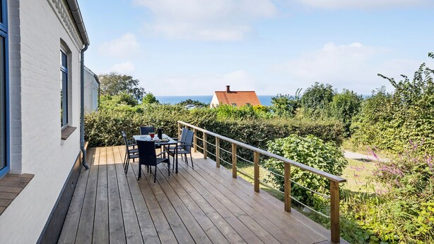 Eine Holzdielen-Terrasse mit Tisch und Stühlen, mit Blick auf einen Garten und das Meer.