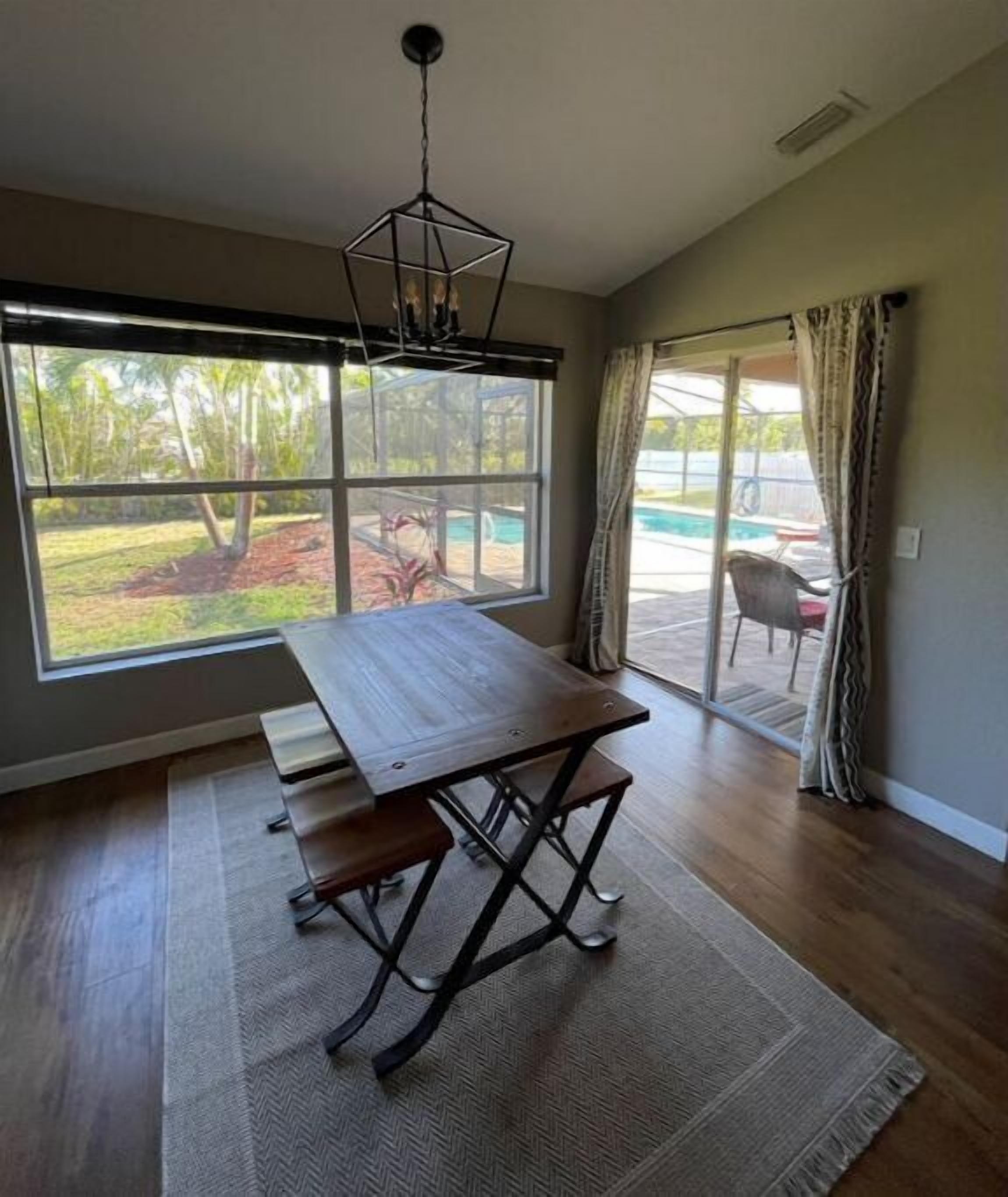 A dining area with a wooden table and benches, large windows with a view of a swimming pool and greenery, and a ceiling light fixture.