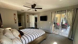 Une chambre à coucher comprenant un lit, un ventilateur de plafond, un téléviseur et une porte-fenêtre coulissante donnant sur un balcon.