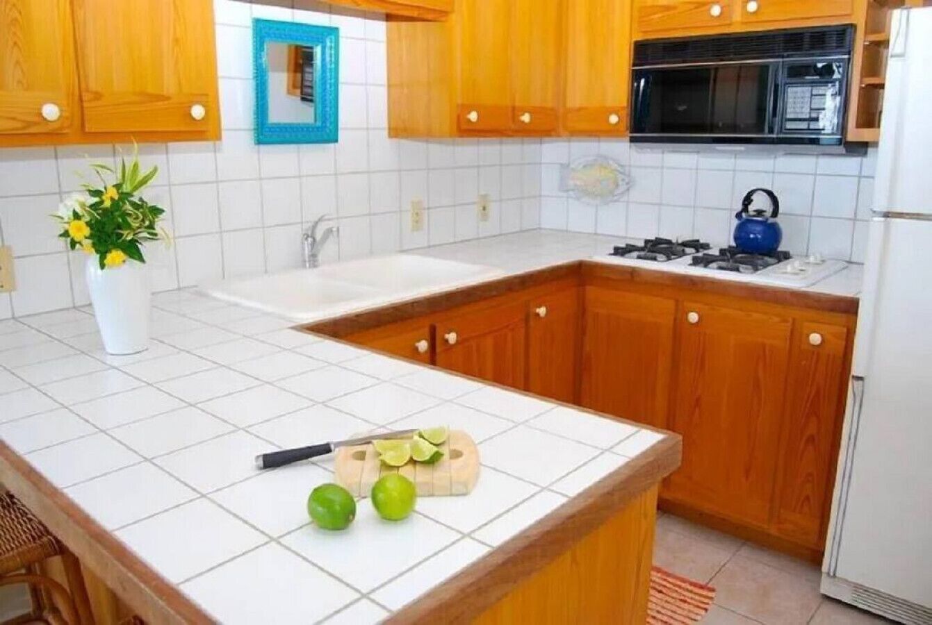 Een keuken met houten kastjes, een witte betegelde spatwand, een fornuis en een magnetron. Op het aanrecht ligt een snijplank met een citroen en een mes.
