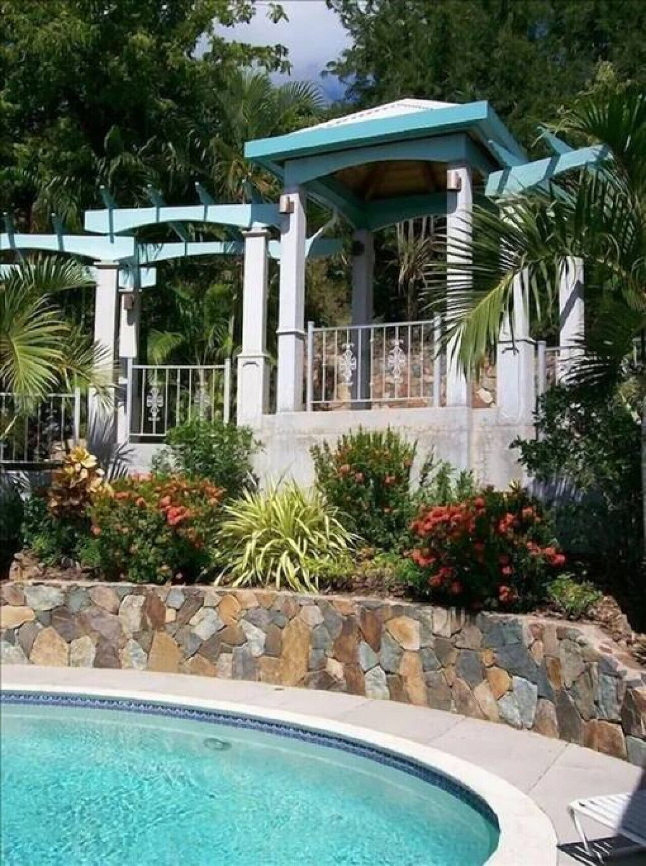 Un área de piscina con muro de piedra, un pabellón y diversas plantas.