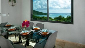 A dining area with a glass table set for six, a vase of flowers, and a view of the sea through a large window. - Tropical Seabreeze- Pool and back up power/solar (Cruz Bay)