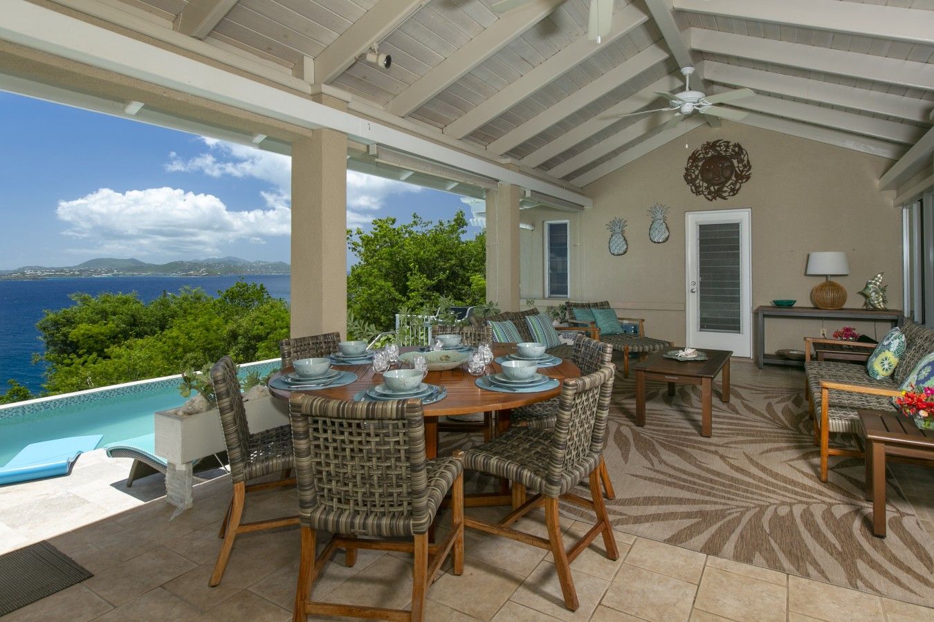 A covered outdoor dining area with a view of the ocean, furnished with wicker chairs and a wooden table set for a meal.
