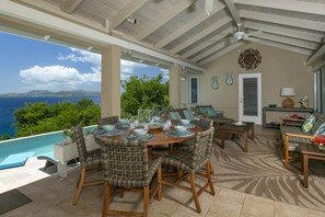 Un espace extérieur abrité, aménagé pour les repas, avec vue sur l’océan, comprenant des chaises en osier et une table en bois dressée pour un repas.