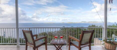 A covered balcony with two wooden chairs and a small table, overlooking a scenic view of the sea and distant land.