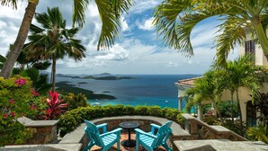 A patio with two blue chairs and a table, overlooking a coastal view with palm trees and a clear sky.