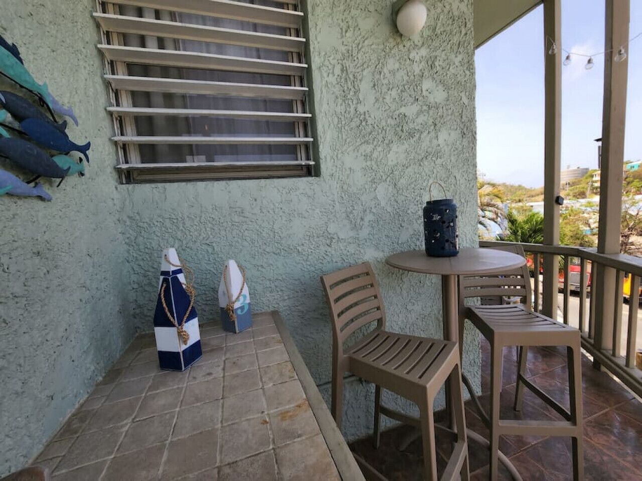 Uma pequena área externa com mesa e cadeiras, uma janela com persianas e um objeto decorativo na parede.