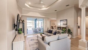 A spacious living room with a light-colored sofa, a coffee table, and a TV mounted on the wall. There is a large window providing natural light and a view of the outdoors. - Dolphins Cove (Cape Coral)
