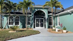 A two-story house with a green exterior, a central arched entrance, and palm trees in the front yard. - Dolphins Cove (Cape Coral)