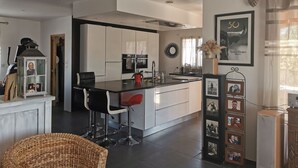 Eine moderne Küche mit weißen Schränken, einer zentralen Kochinsel und Barhocker. An der Wand hängt eine Collage gerahmter Fotos und links steht ein dekorativer Laternenleuchter.