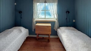 Et værelse med to enkeltsenge, en træbænk, et vindue med blonde gardiner og udsigt til havet.