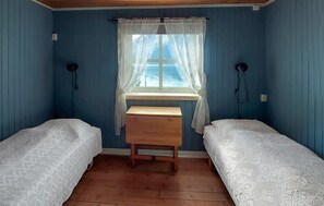 A room with two single beds, a wooden bench, a window with lace curtains, and a view of the sea.