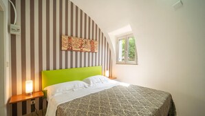 Ein Schlafzimmer mit gestreiftem Wandpapier, einem grünen Kopfteil, einer gemusterten Bettdecke, einer Nachttischlampe und einem Fenster mit Blick ins Grüne.