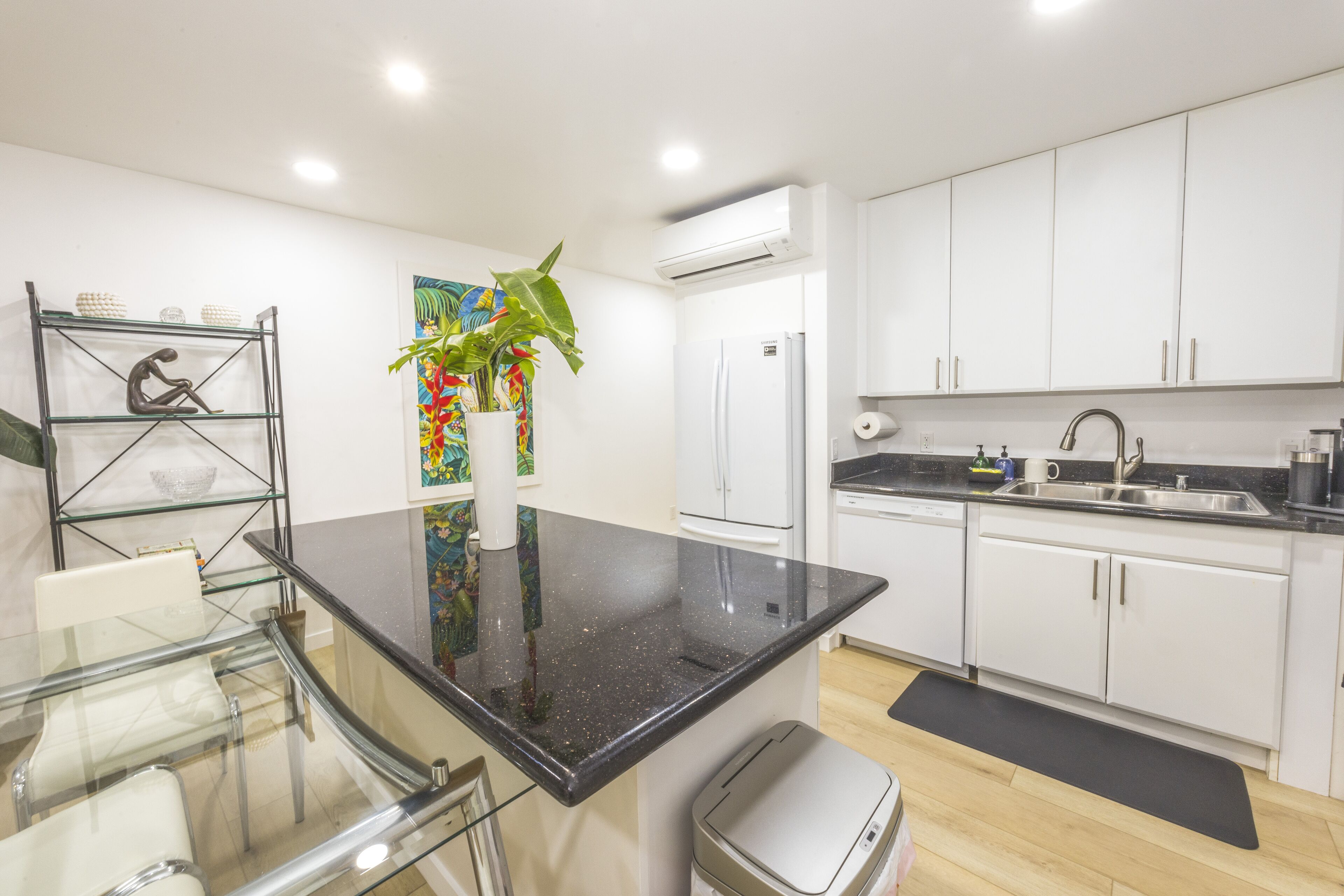 Una cocina moderna con armarios blancos, encimera negra, mesa de comedor de vidrio y una planta en maceta.