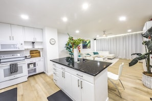 A modern kitchen with white cabinetry, a black countertop, and a microwave. There is a dining area with a white table and chairs, and a living room with a white sofa and a plant.