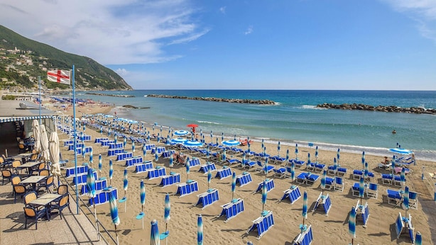 Une plage bordée de rangées de transats et de parasols bleus, un drapeau et une digue rocheuse en arrière-plan.
