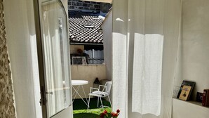 Ett rum med en vit soffa, ett litet runt bord med en vas med blommor och utsikt över en trädgård genom en öppen dörr.