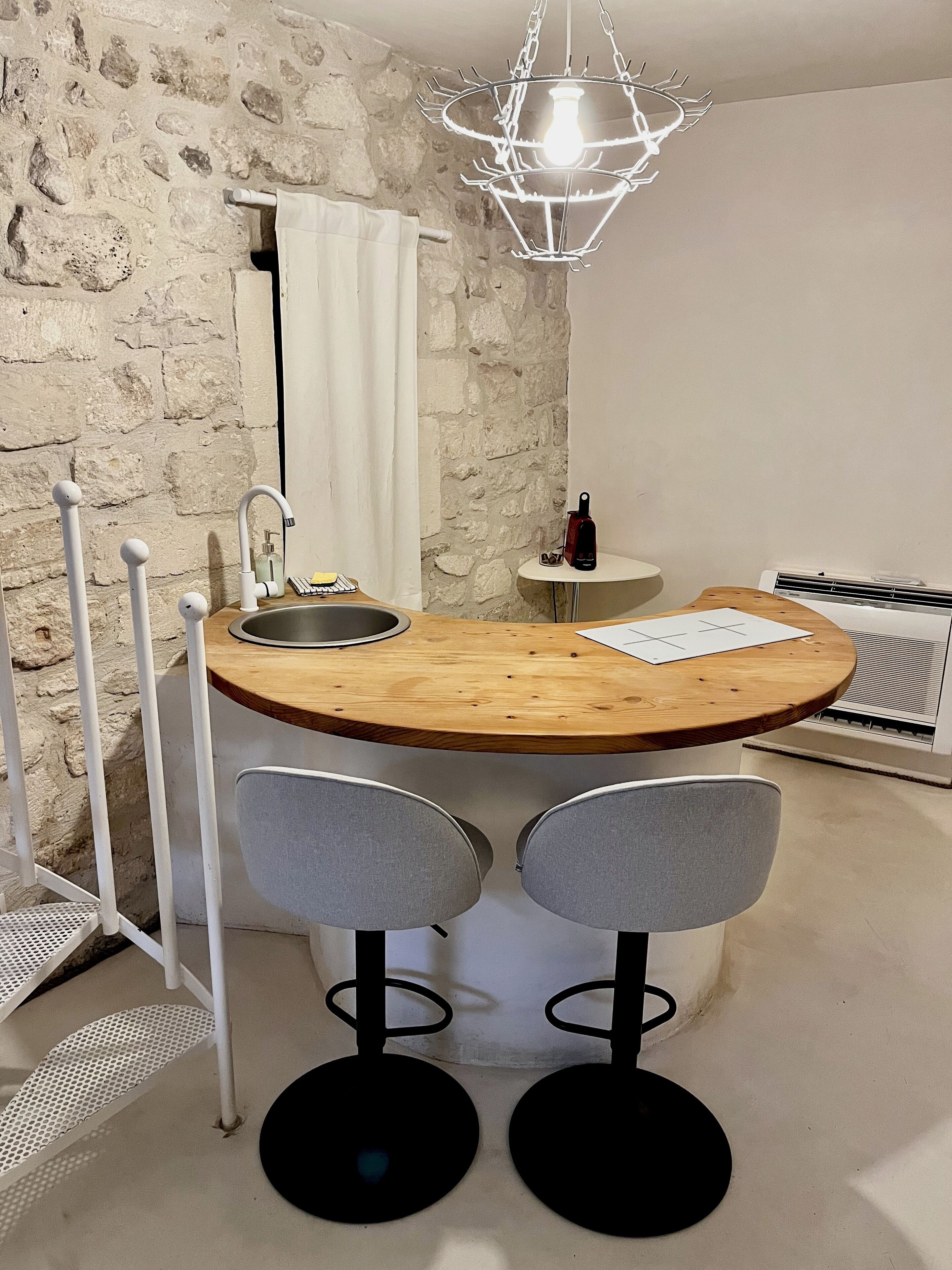 A compact kitchen with a stone wall, a wooden dining table with two chairs, a hanging light fixture, and a built-in sink.