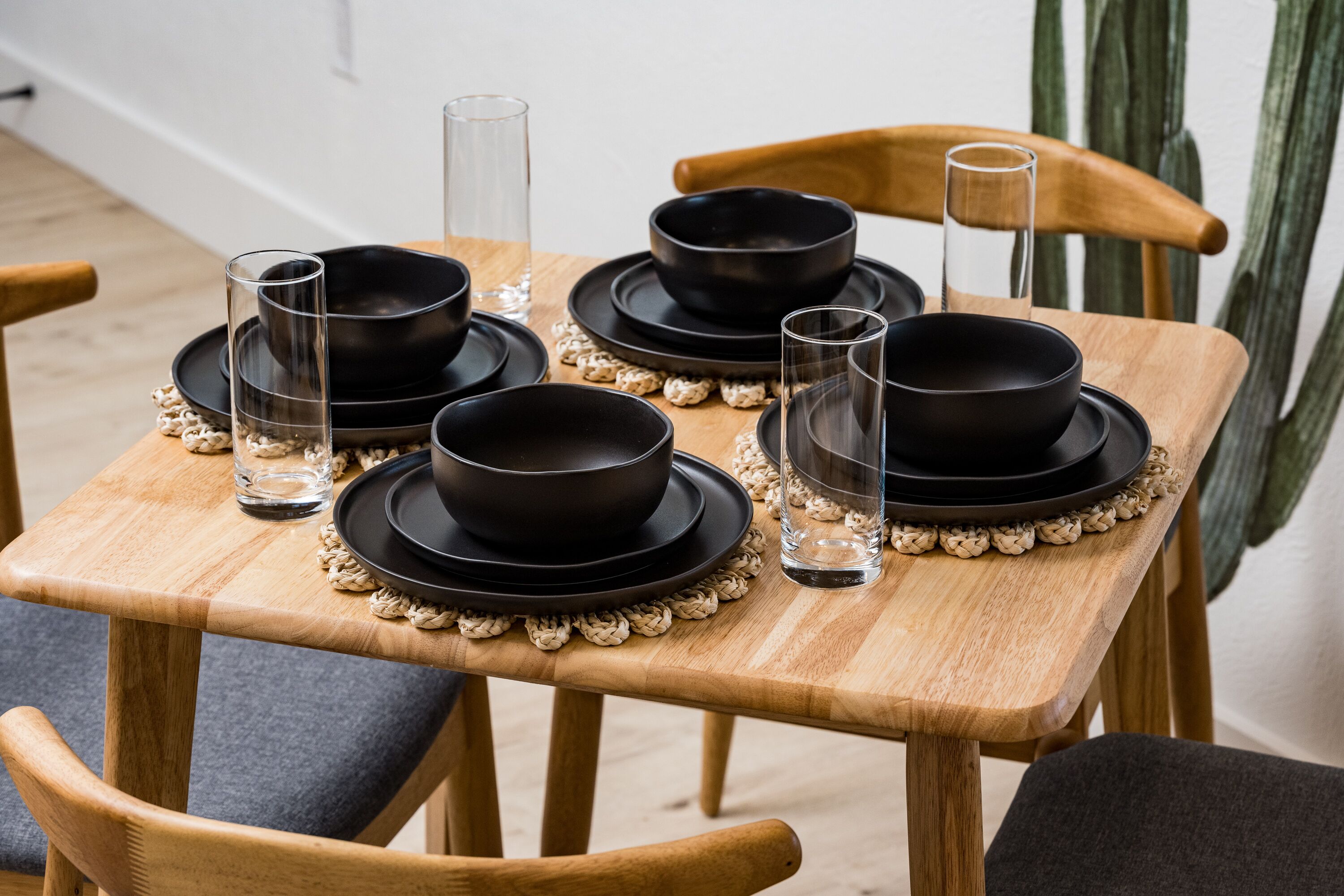 A wooden dining table set with black bowls, plates, and clear glasses, surrounded by wooden chairs.