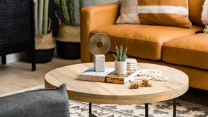 Uma mesa de centro redonda de madeira com livros, um objeto decorativo de metal, uma pequena planta em vaso e um fio de contas brancas.