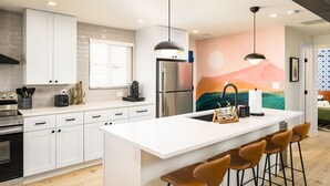 Uma cozinha moderna com armários brancos, uma ilha central com banquetas, eletrodomésticos de aço inoxidável e iluminação pendente.