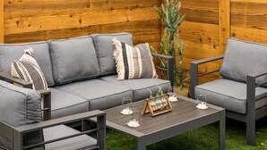 Uma área de estar moderna ao ar livre, com um sofá cinza e poltronas combinando, uma mesa de centro de madeira e uma planta de cacto.