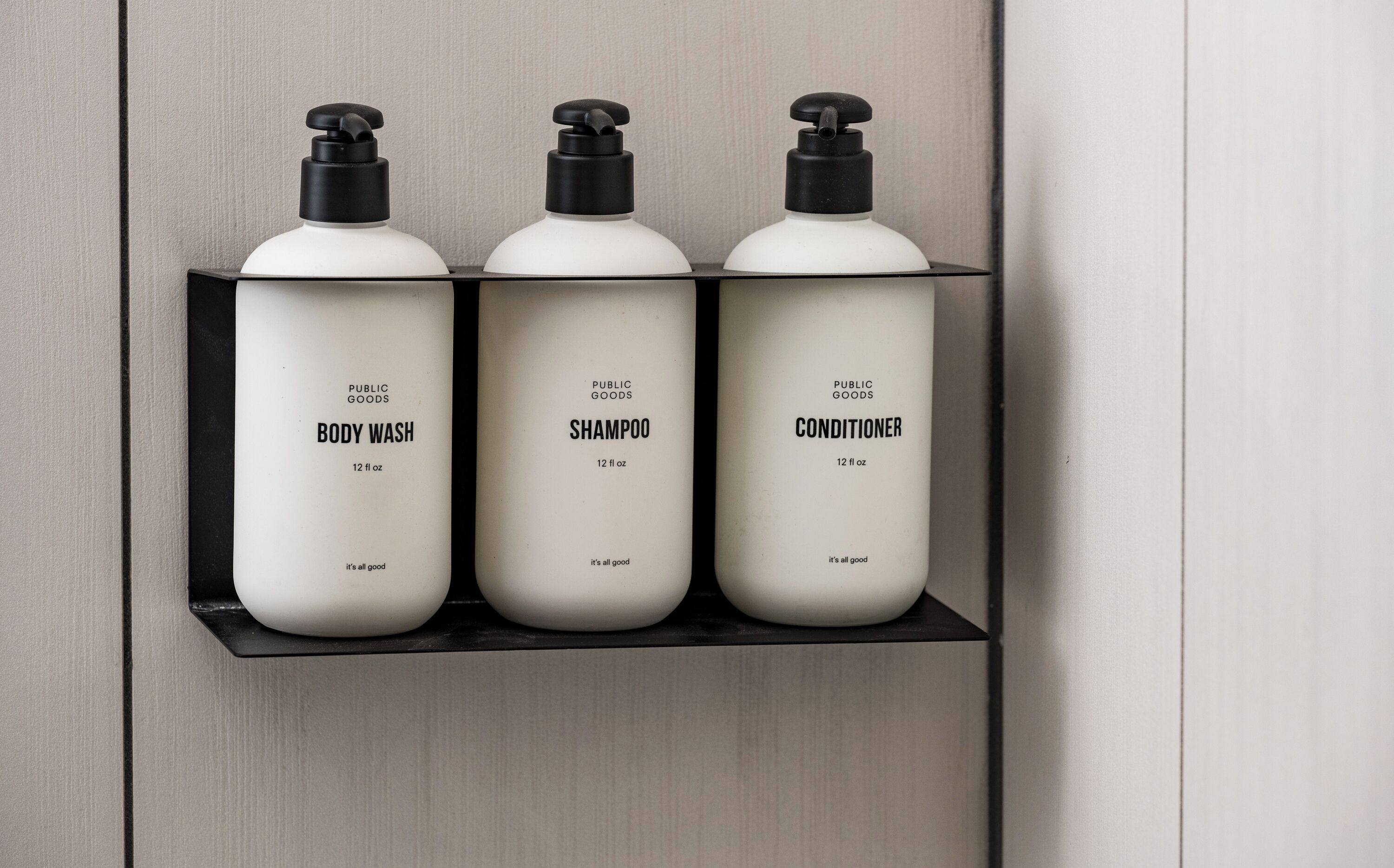 Tres botellas de jabón de cuerpo, champú y acondicionador sobre un estante negro frente a una pared.
